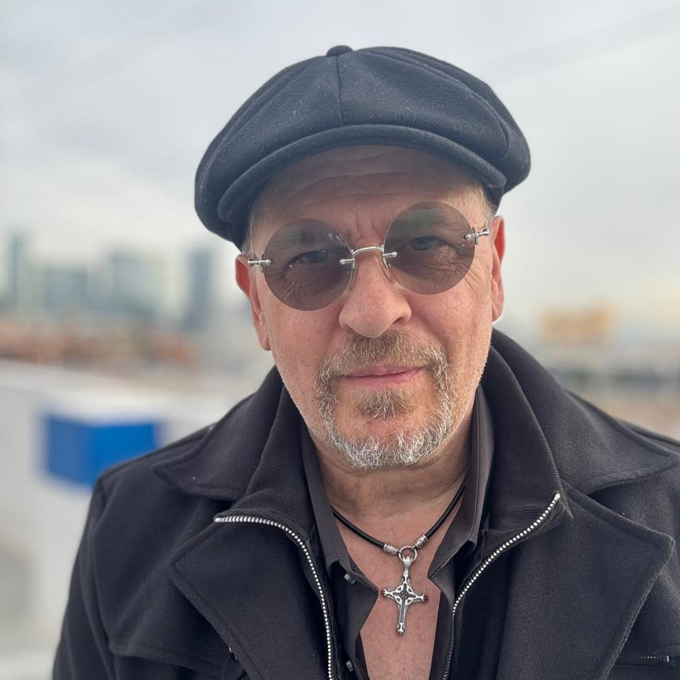 Photo of Wayne Wallace with Black News Boy Hat and round no rim glasses and black coat with cross and goatee and mustache facial hair